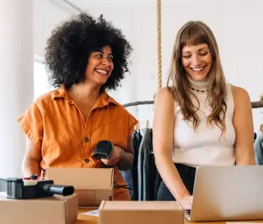 Black woman with an afro wearing a orange romper talking to her business partner, a blonde woman wearing a white sleeveless shirt talking about setting up their registered agent with LegalZoom as they mail packages to customers.