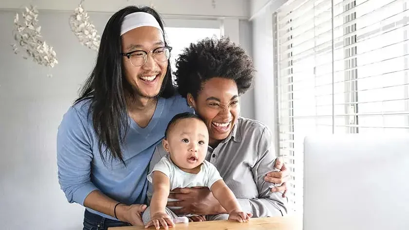Two parents hold their child at a desk, all three laughing at the computer