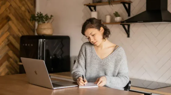 A woman stands at a desk talking on her phone while taking notes on a notepad to the side of her laptop.