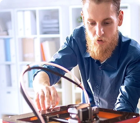 White man with a red beard and a blue shirt checks a three d printer