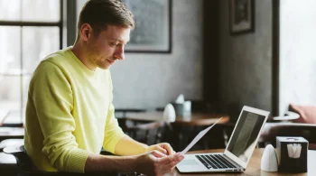 Man in yellow sweater reading papers at a laptop