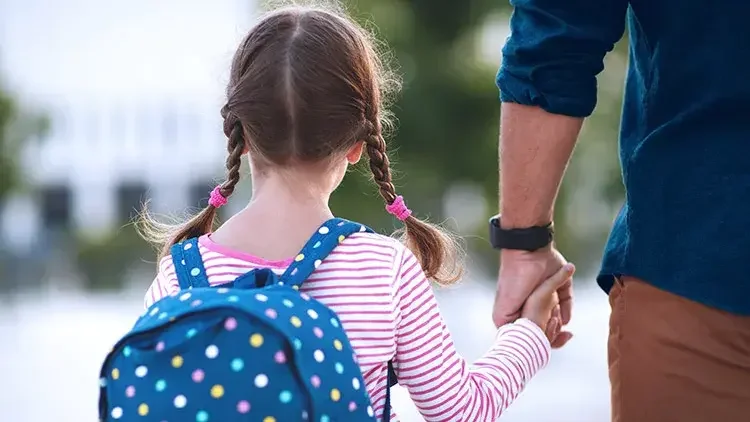 Dad's hand holds young girl in pink shirt and blue backpack's hand.