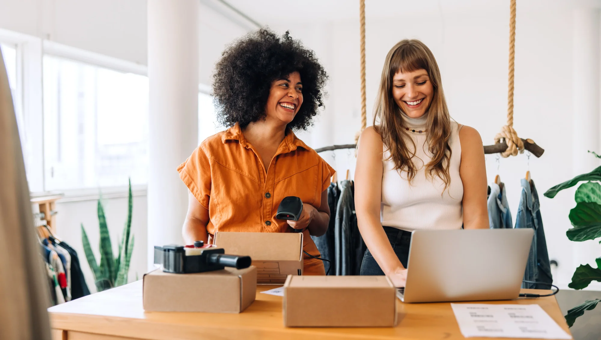 two women smile and laugh standing over a table with boxes and a laptop