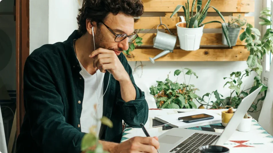 Man wearing glasses and earbuds writes in his journal next to his laptop.