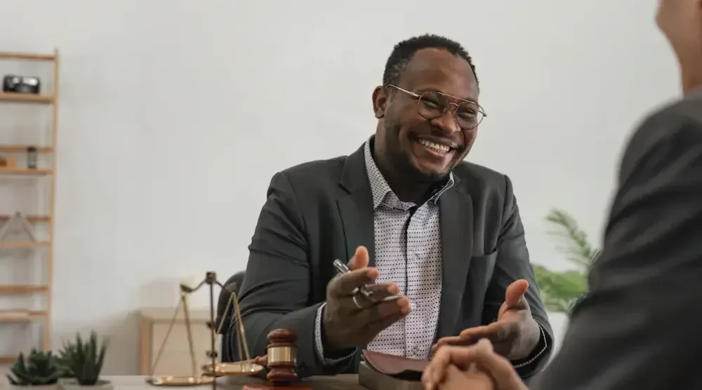 An estate planning attorney, seated behind a desk, shares a laugh with a client as he explains the differences between a will and a trust and which might work better for him.