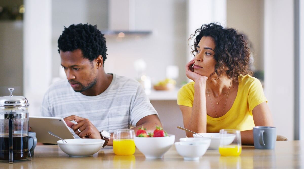 A man and woman sit in the kitchen with breakfast in front of them. The woman looks over at the man while he scrolls on his phone.