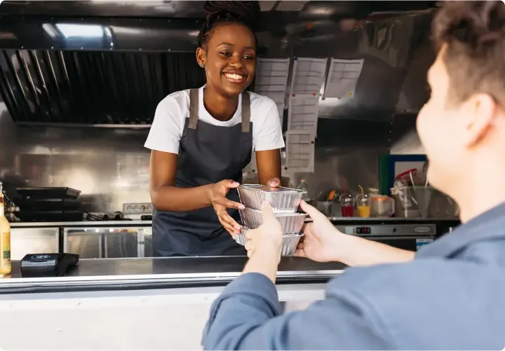 a young woman works the counter of a food truck and hands an order to a customer.