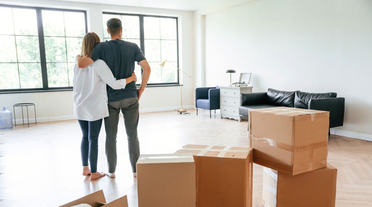 A man and a woman embrace in a bare living room next to a stack of moving boxes