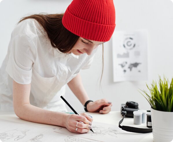 Woman in a shirt and red beanie writes with a pencil on her desk.