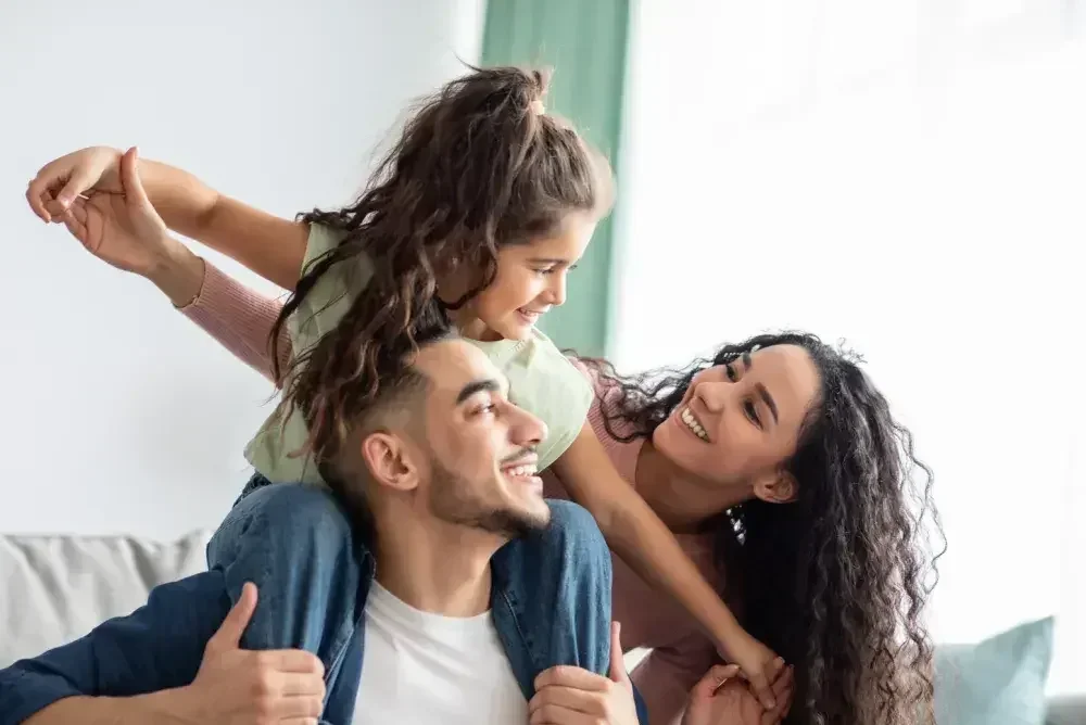 A happy mother, father, and daughter smiling and playing together, with the daughter sitting on her father's shoulders in a bright living room.
