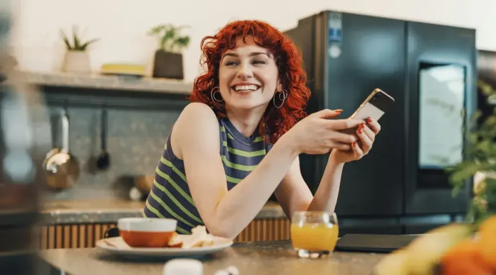 A redhead barista stands smiling behind the bar at her cafe.