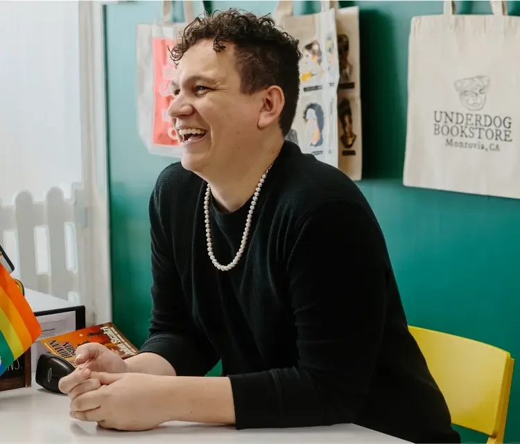 A man wearing a black shirt with a necklace is laughing at his desk.