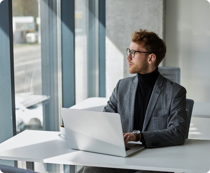 White man with red hair, glasses and a 5 o'clock shadow  wearing a grey suit with a black turtle neck sitting in the office with a white laptop filling out his business dissolution with LegalZoom.