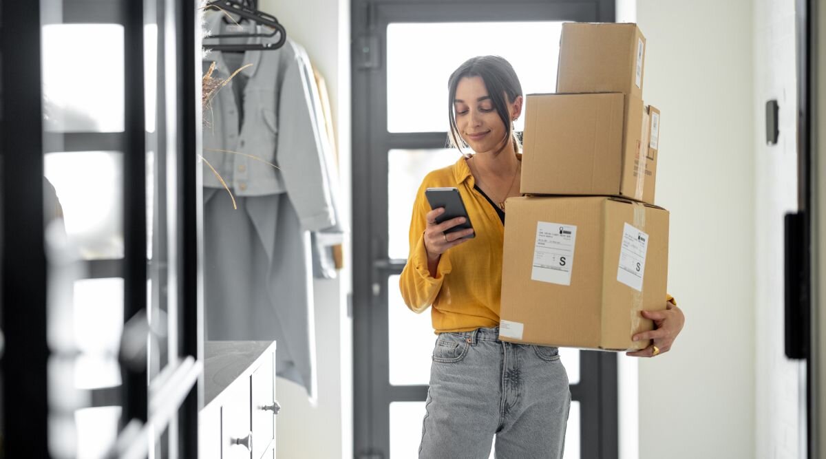 A woman stands in the doorway holding a stack of packages.