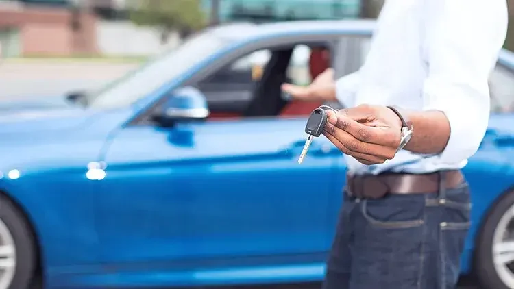 A man stands in front a of a blue sedan, handing the viewer the key