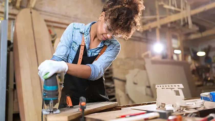 Woman in a denim shirt and a black apron using a power tool to cut a piece of wood for a sign for her business.