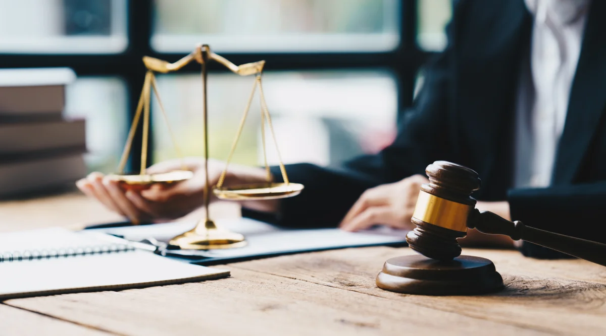 A judge sits at a desk as she balances a "scale of justice" in one hand. Beside her is a gavel resting on a block.