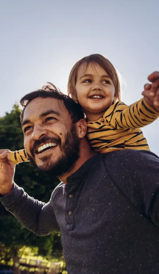 A smiling father carries his daughter on his shoulders. The daughter is spreading her arms out in flight.