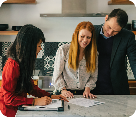 A real estate agent sitting at a kitchen counter with a smiling couple, reviewing and signing property documents together.