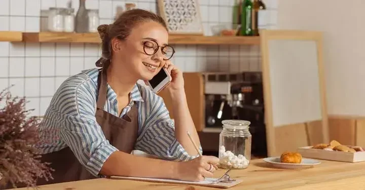 A small business owner sitting in her coffee shop, talking on the phone and writing on a clipboard.