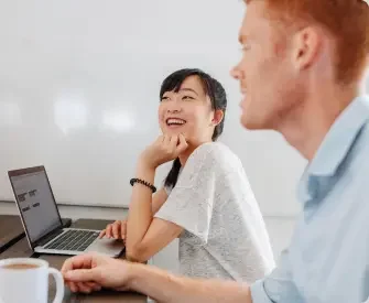 An asian woman in the background sits at her computer and laughs with a man in the foreground.