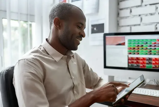 An African American man wearing a collared shirt sits at his desk and uses a tablet.