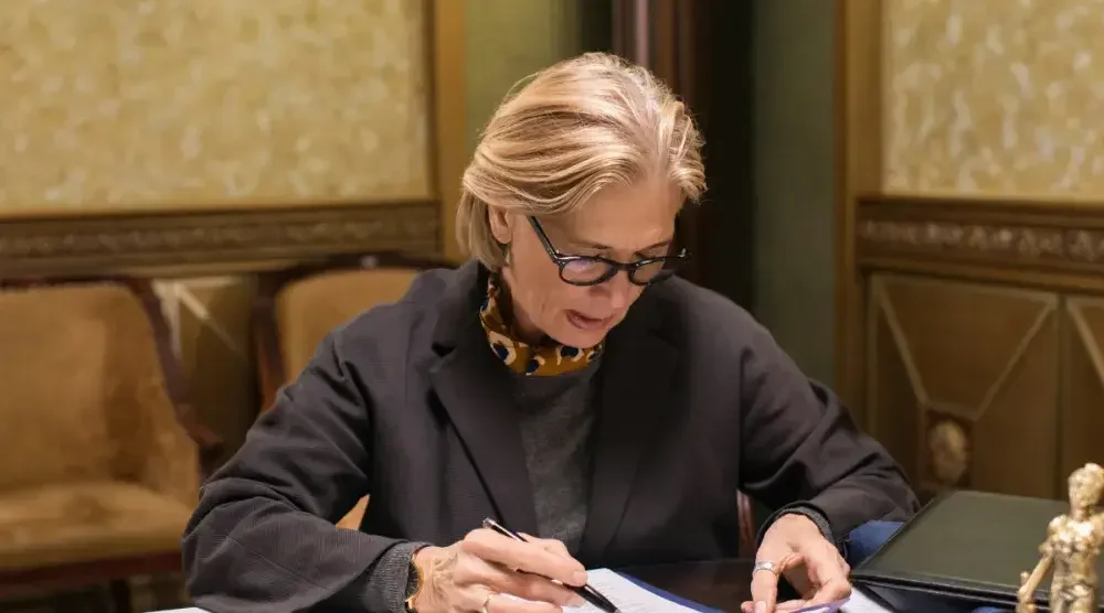 A blonde woman with glasses sits at her desk taking notes on paper.