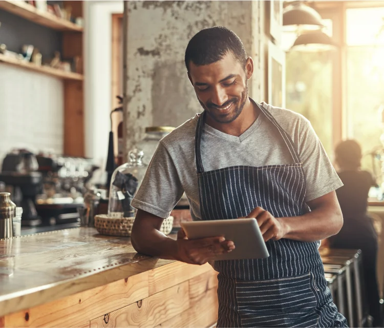 A man in an apron smiles at this ipad, while leaning on a wooden desk.