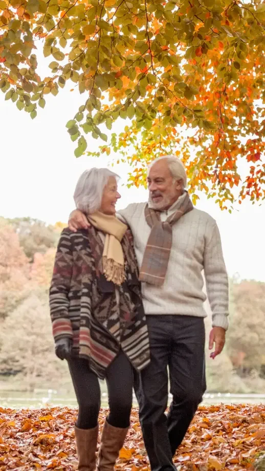 An older man and woman smile into each other's eyes as they walk through orange and red leaves in the fall.
