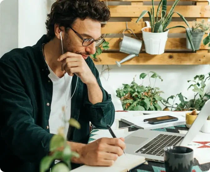 Man wearing glasses and earbuds writes in his journal next to his laptop.