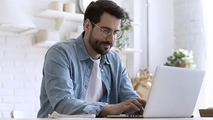 Young man in cozy white kitchen with a book and flower vase nearby, uses his laptop to research how to get a Florida business license.