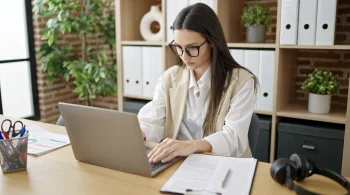 Woman with glasses typing on a laptop in an office