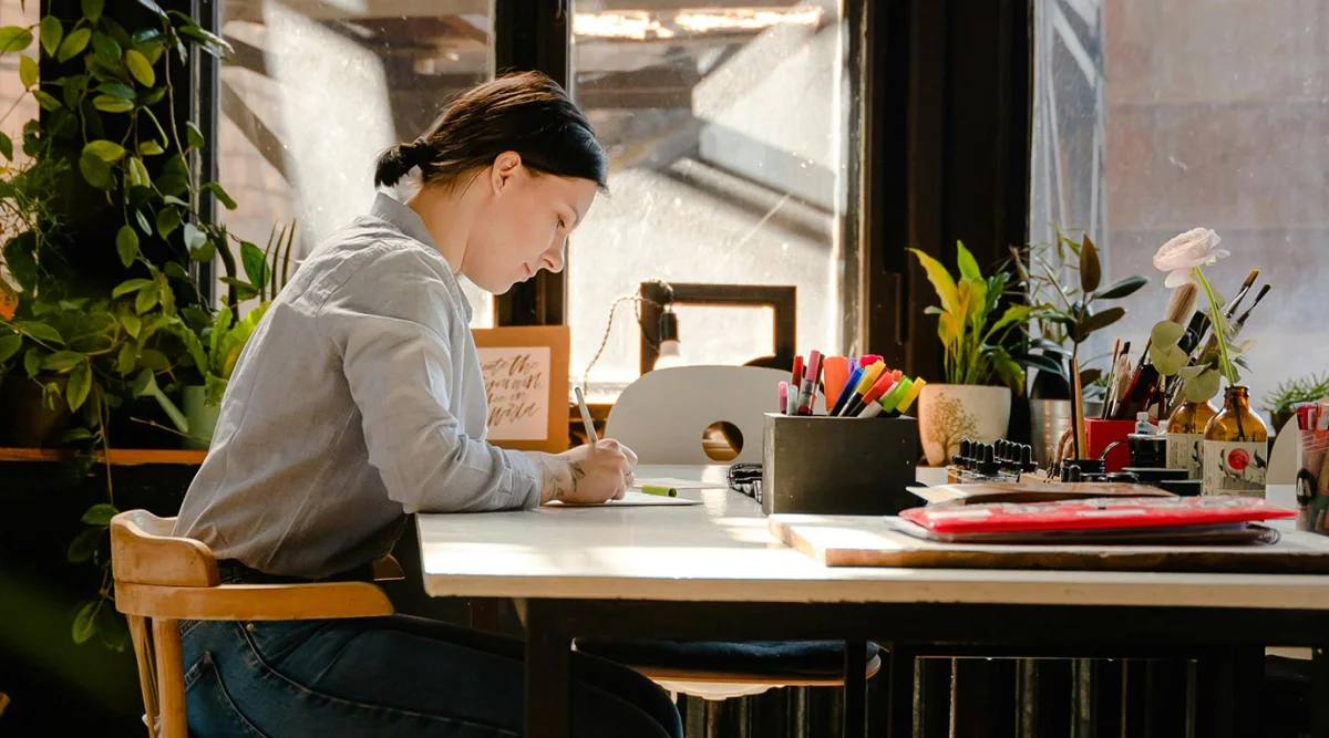 A woman sits at a desk covered in art supplies