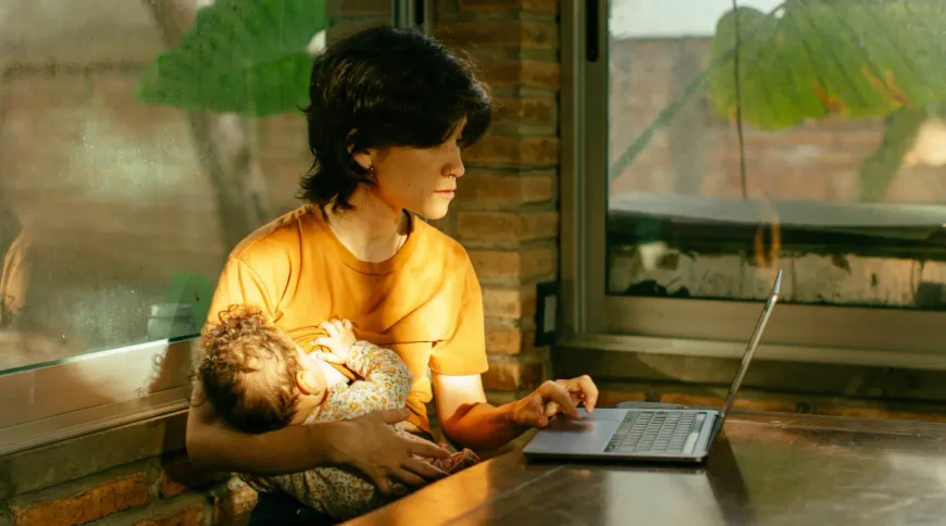 A woman breastfeeds while researching trusts on her laptop.