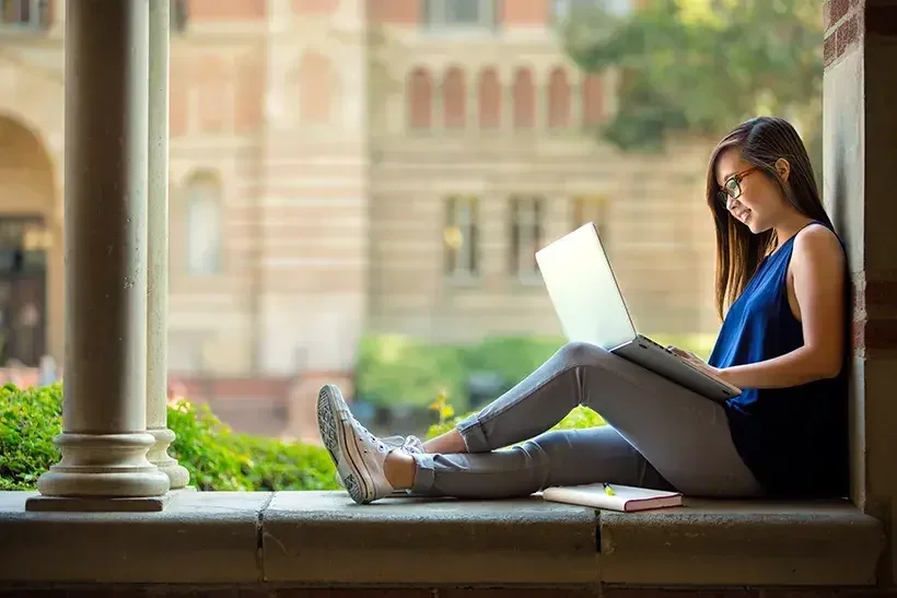 A young adult on the college premises is working on an assignment on her laptop.