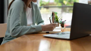 Person writing notes with a laptop on a wooden table