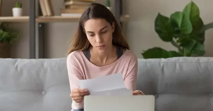 A woman sits on a living room couch and reads a letter. 