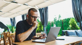 Man with a drink using a laptop outdoors under a canopy