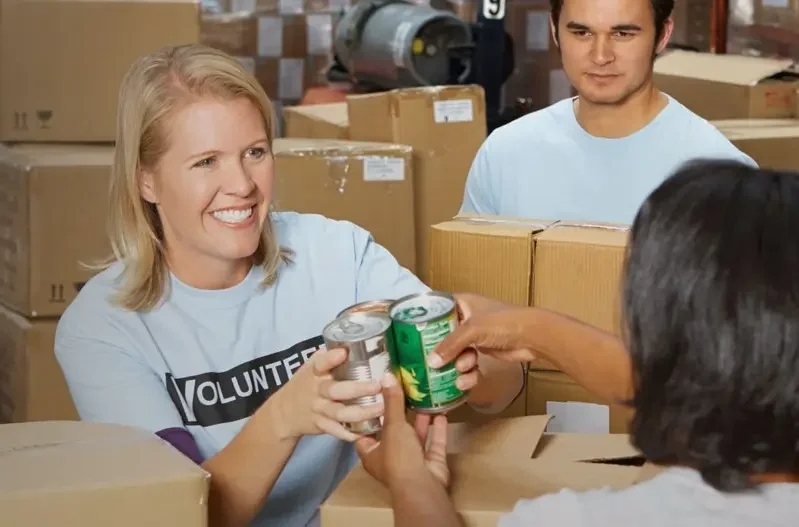 Volunteers in a warehouse distribute canned food, with a smiling woman handing cans to a recipient amid stacked boxes.