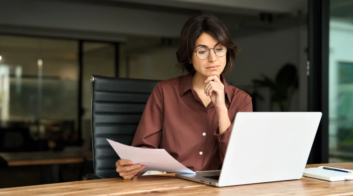 Thoughtful person holding papers next to a laptop