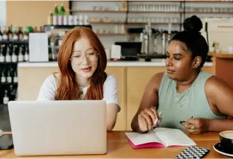 two women study a laptop while one takes notes on paper.