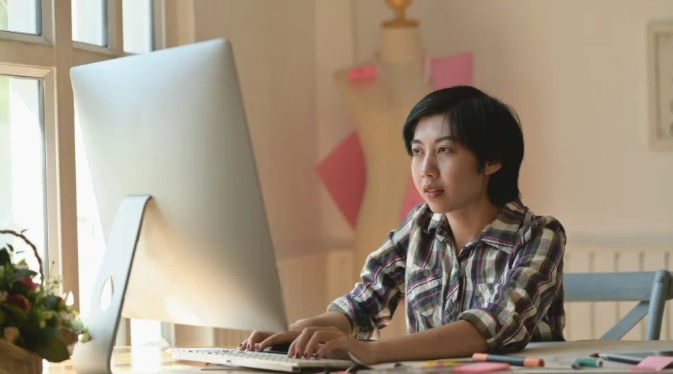 A woman sits at a desk in an office working on a desktop computer.