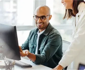 man wearing glasses smiles at his desktop, and speaks to his coworker.