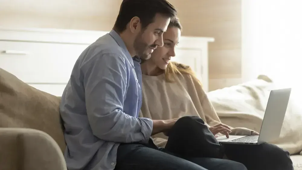 A smiling couple is sitting on their sofa and reading about the basics of 'I Love You' wills on their laptop. 