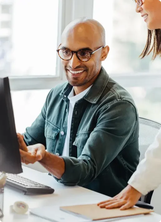 man wearing glasses smiles at his desktop, and speaks to his coworker.