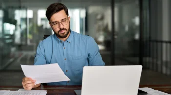 Man in blue shirt reviewing a paper at a laptop