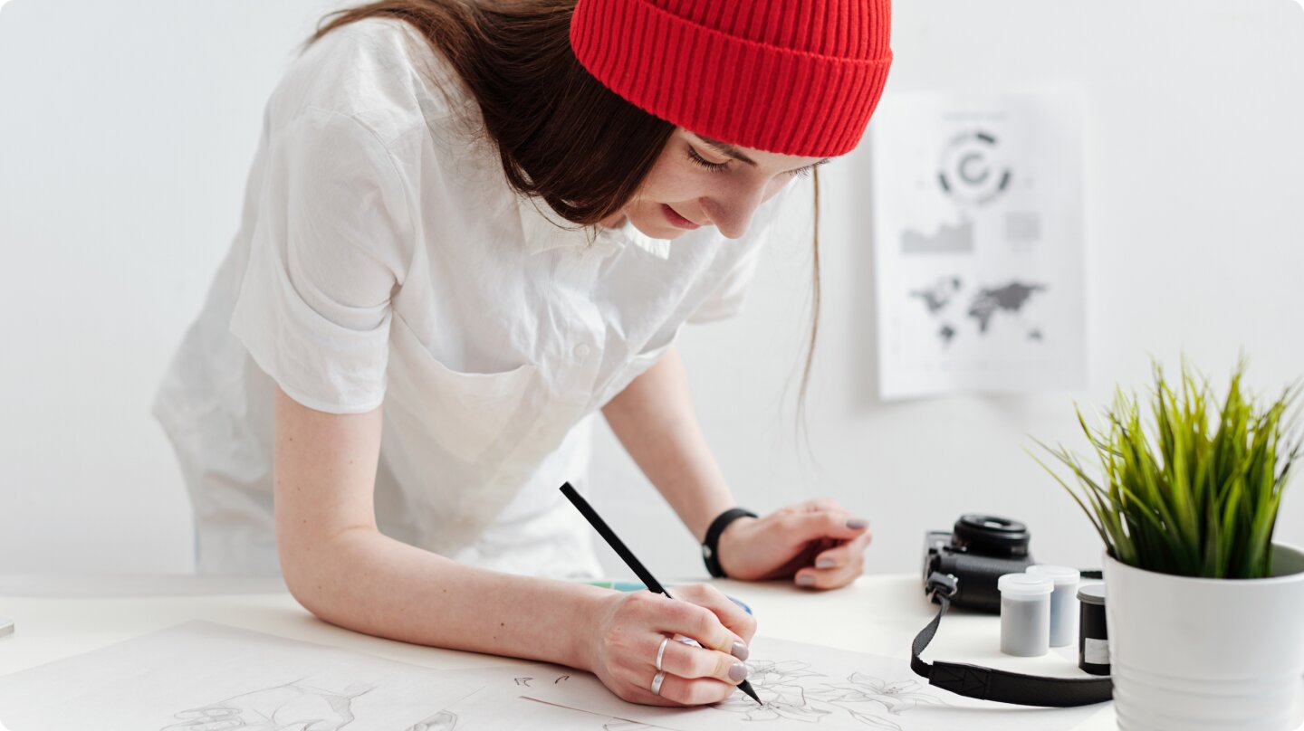 Woman in a shirt and red beanie writes with a pencil on her desk.
