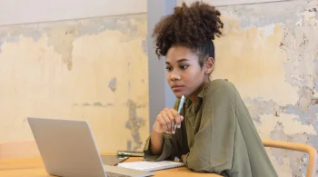 Woman pondering with a pen, looking at a laptop in a rustic room