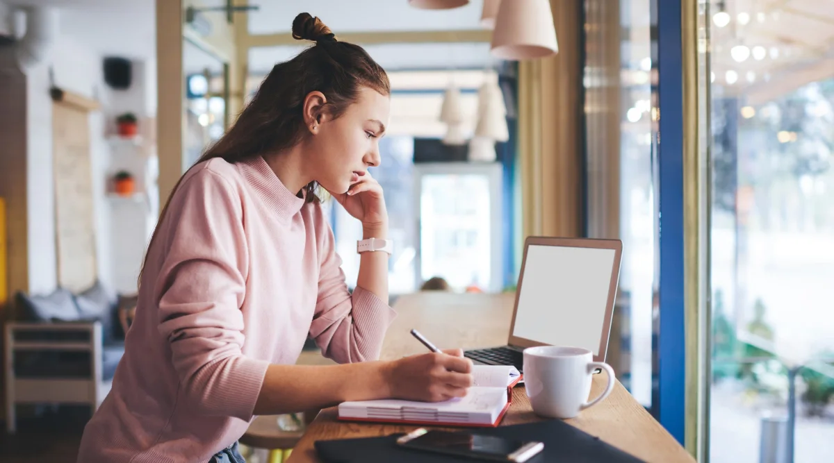 A woman taking notes while looking at her laptop.