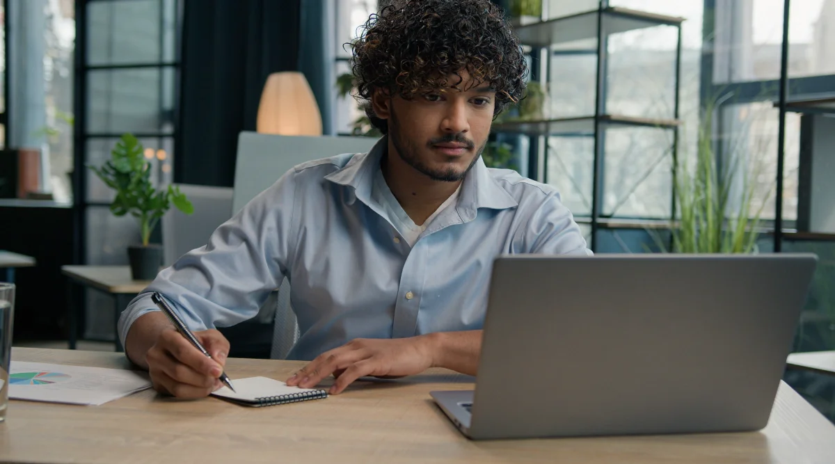 Person writing notes while using a laptop at a desk